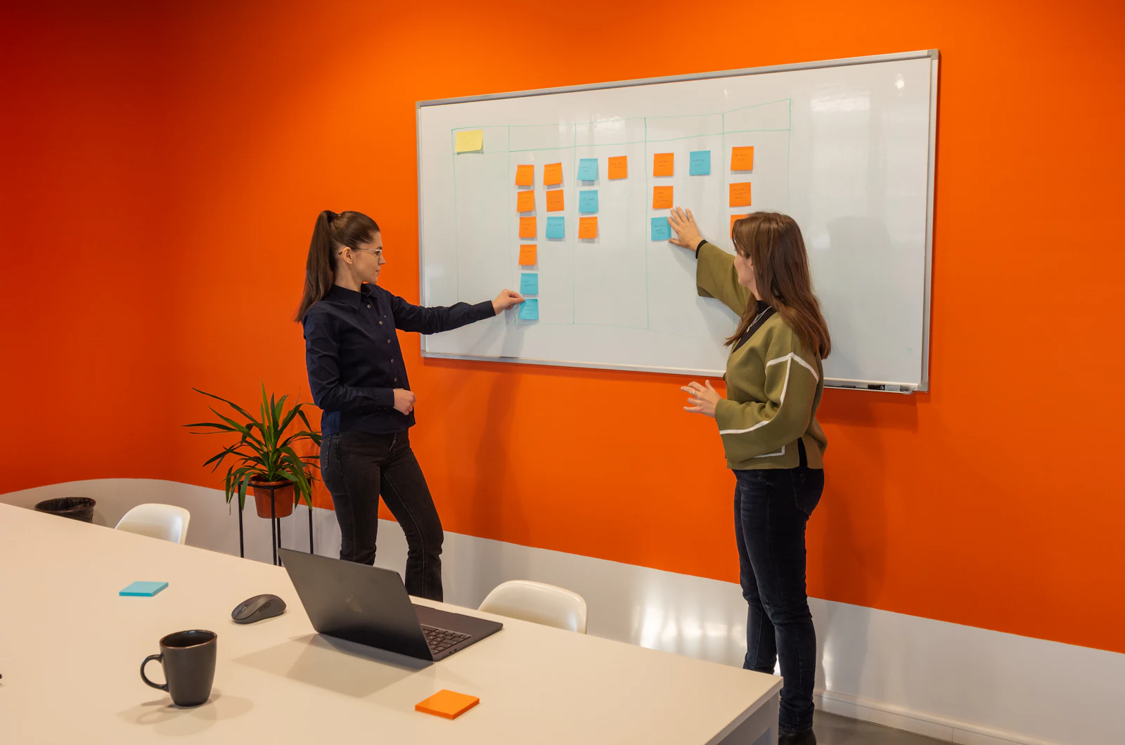Twee collega's plannen het werk op een whiteboard voor een oranje wand